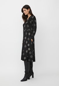 Robe noire à manches longues, décolleté en V, ornée de motifs géométriques argentés. Le tissu semble lisse, avec des poches et une coupe au genou.