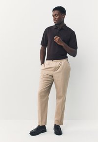 Dunkelbraunes Poloshirt kombiniert mit beigefarbenen Faltenhosen und schwarzen Loafers, präsentiert vor hellem Hintergrund. Einfaches Design und unifarbene Töne.