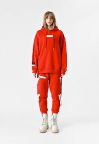 Sudadera roja extragrande con un bolsillo frontal, combinada con joggers a juego que presentan patrones bordados en blanco. Termina con botas blancas con cordones.