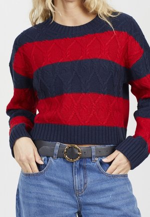 Femme portant un pull en maille à larges rayures bleu marine et rouge, un jean bleu et une ceinture noire avec une boucle ronde dorée.