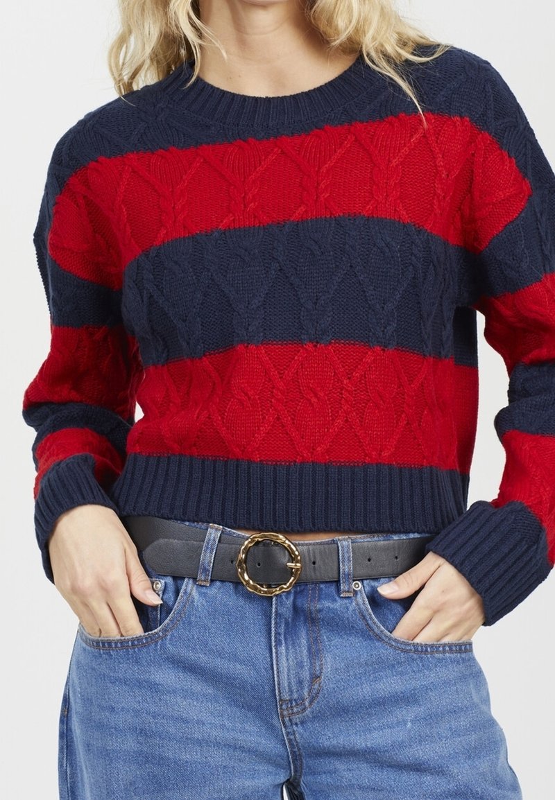 Femme portant un pull en maille à larges rayures bleu marine et rouge, un jean bleu et une ceinture noire avec une boucle ronde dorée.
