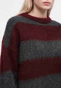 Personne portant un pull duveteux avec de larges rayures horizontales grenat et grises, montrant le haut du torse et le cou.