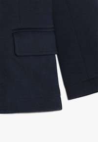 Tkanina marynarki w ciemnym granatowym kolorze o fakturowanym wykończeniu, z klapką kieszeni po lewej stronie i starannym przeszyciem wzdłuż krawędzi.