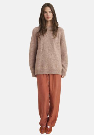 Maglione di lana rosa chiaro con una vestibilità rilassata, abbinato a pantaloni fluidi color ruggine. Il modello indossa scarpe basse con accenti viola.