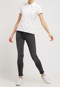 Žena v bílé polo košili s malými růžovými potisky tenisových raket, černých skinny džínech a bílých teniskách, stojící s rozpaženýma rukama.