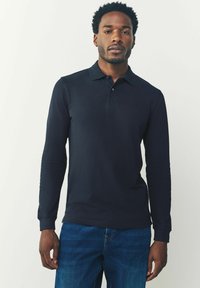 Camisa polo de manga larga en color navy con cuello y un discreto detalle de logo en el pecho, combinada con jeans azules. Textura suave y lisa.