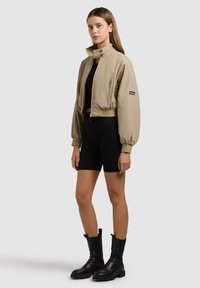 Femme debout portant une veste courte beige, un crop top noir, un short de cycliste noir et des bottes noires mi-mollet sur un fond uni.