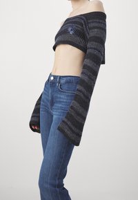 Harmaan raidallinen lyhyt neulepaita, jossa on olkaimia paljastava muotoilu, ribbitekstuuri ja pitkät, levenevät hihat, yhdistettyinä istuviin sinisiin farkkuihin.