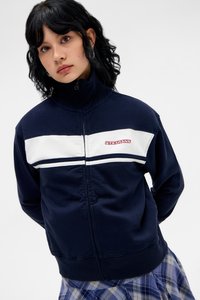 Dunkelblaues Zip-Up-Sweatshirt mit weißen horizontalen Streifen und rotem Logo. Mit hohem Kragen und strukturiertem Stoff. Kombiniert mit einem karierten Rock.