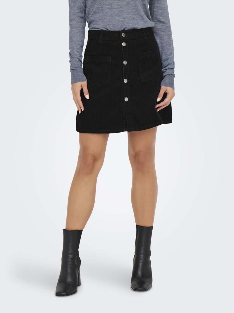 Donna che indossa una minigonna nera in velluto a costine con bottoni sul davanti e tasche frontali, una maglia grigia a maniche lunghe e stivaletti neri con tacco.