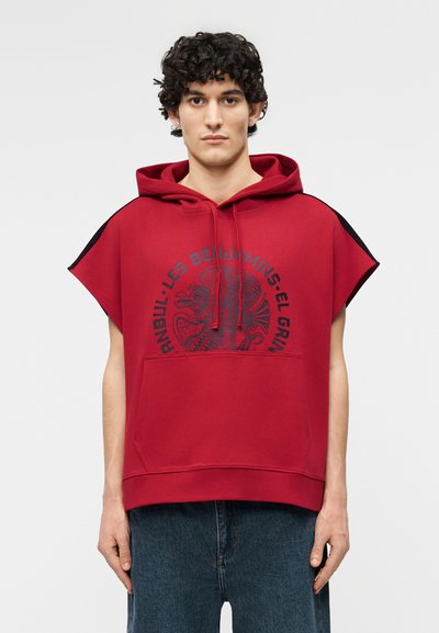 Jongere met krullend haar, gekleed in een rood mouwloos hoodie met een zak aan de voorkant en zwarte tekst en illustratie op de borst.