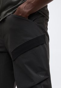 Κοντινό ενός ατόμου που φοράει μαύρο παντελόνι με το χέρι στην μπροστινή τσέπη και διακριτικό λογότυπο στο μηρό.