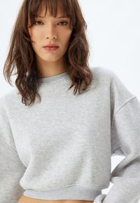Sudadera gris corta con cuello redondo, mangas largas y puños acanalados. La tela parece suave con una sutil textura jaspeada.