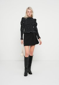 Bluză neagră cu buline și mâneci bufante, asortată cu o fustă mini neagră și cizme negre până la genunchi. Modelul ține o poșetă mică albă.