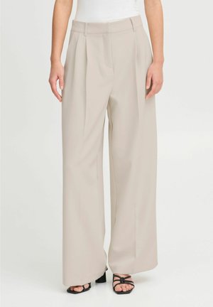 Pantalon beige à jambes larges avec plis à l'avant, porté avec des talons noirs à brides ouvertes et un haut blanc, montré de la taille aux pieds.