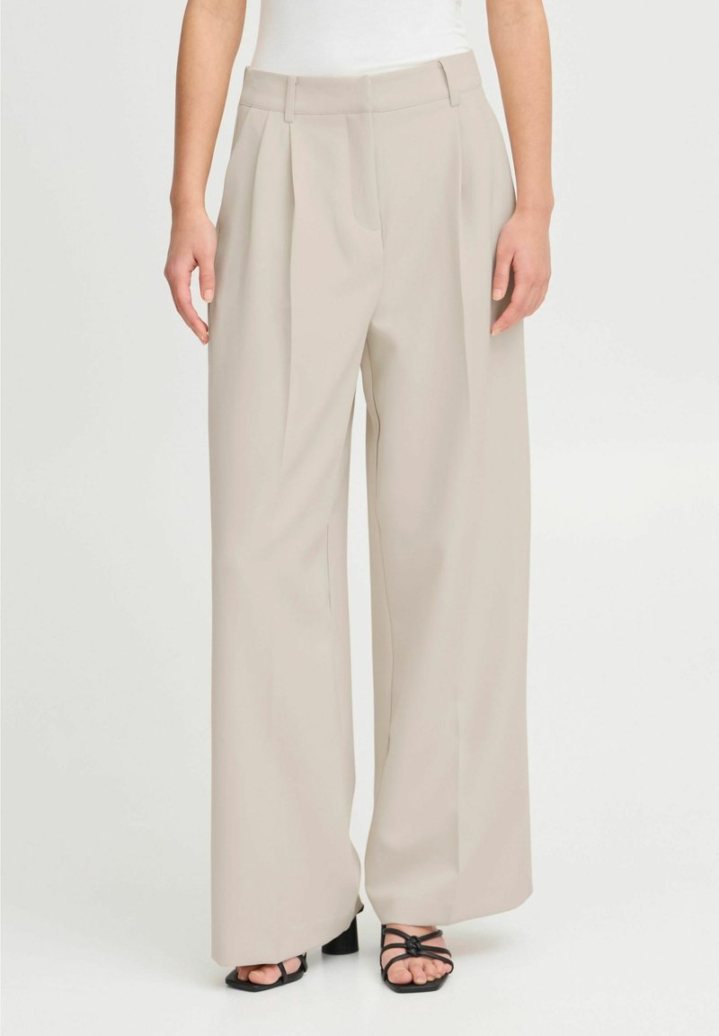 Pantalon beige à jambes larges avec plis à l'avant, porté avec des talons noirs à brides ouvertes et un haut blanc, montré de la taille aux pieds.