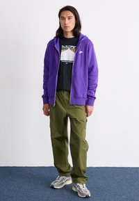 Un hanorac cu fermoar violet peste un tricou negru cu grafică, combinat cu pantaloni cargo verzi măslin și pantofi de alergare gri cu detalii din plasă.
