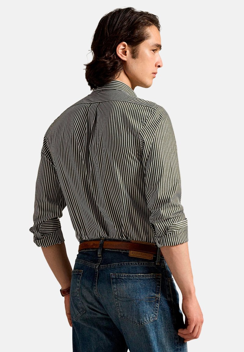 Chemise à manches longues rayée noir et blanc, avec un col classique. Le modèle porte un jean bleu, manches retroussées, montrant la texture du tissu.