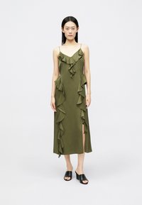 Mulher vestindo um vestido midi verde-oliva com babados e alças finas, combinado com sandálias de salto alto aberto preto, em pé sobre um fundo branco.