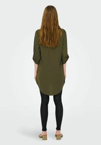 Una lunga camicia verde oliva con orlo arrotondato e maniche arrotolate, abbinata a leggings neri aderenti e sandali con tacco quadrato color beige.