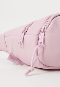 Sac banane en tissu rose avec surface texturée, compartiments à double fermeture éclair et tirettes assorties. Présente une petite étiquette de logo.