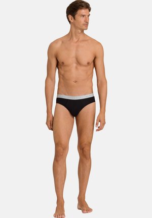 Mann steht barfuß, trägt schwarze Slips mit einem hellgrauen Bund und schaut seitlich vor einem einfarbigen Hintergrund.