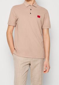 Beżowa koszulka polo z krótkimi rękawami, dwoma czarnymi guzikami i czerwoną metką marki na piersi. Miękki materiał bawełniany. Luźny krój.