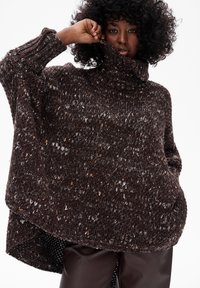 Kobieta z kręconymi włosami, ubrana w luźny, cętkowany, ciemnobrązowy sweter z golfem oraz ciemne skórzane spodnie, na jednolitym białym tle.