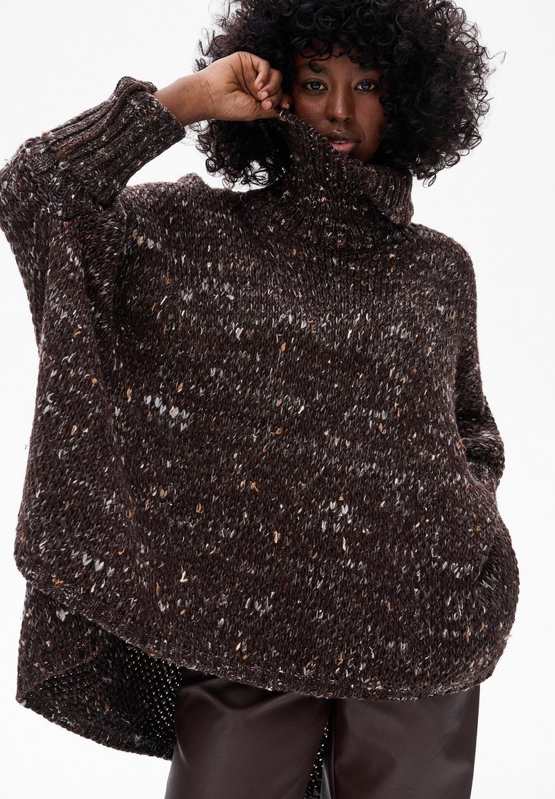 Kobieta z kręconymi włosami, ubrana w luźny, cętkowany, ciemnobrązowy sweter z golfem oraz ciemne skórzane spodnie, na jednolitym białym tle.