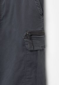 Pantalonii cargo gri închis au o buzunar lateral cu fermoar, un material texturat și coasere întărită pe cusături.