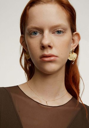 Pendientes de oro con un patrón ondulado y una cuenta transparente colgante, llevados con una blusa marrón transparente y un delicado collar de oro, que resaltan un diseño intrincado.