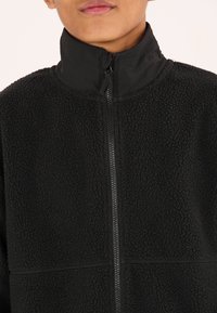 Veste en polaire noire avec col montant et fermeture éclair. Présente un tissu texturé, des accents lisses et un design structuré.