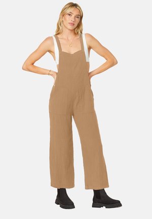 Tan linen overalls with wide legs, adjustable shoulder straps, and two front pockets. Paired with black ankle boots.