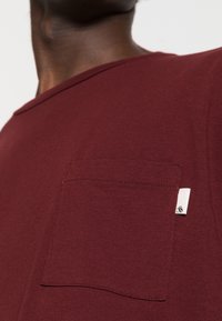 Primo piano di una persona che indossa una maglietta color marsala con una tasca sul petto cucita e un piccolo etichetta bianca sul lato destro.