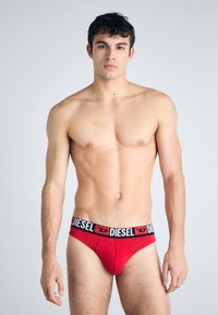 Κόκκινοι σλιπ με μαύρη ζώνη που διαθέτει το λογότυπο "DIESEL". Λείο ύφασμα, άνετη εφαρμογή και μινιμαλιστικός σχεδιασμός. Το μοντέλο εκτίθεται σε ουδέτερο φόντο.