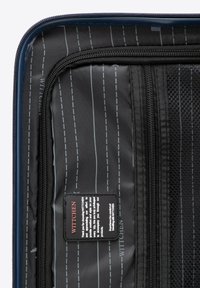 Interieur van een blauwe harde koffer met zwart stof en grijze strepen, ritscompartimenten en een merklabel met verzorgingsinstructies.