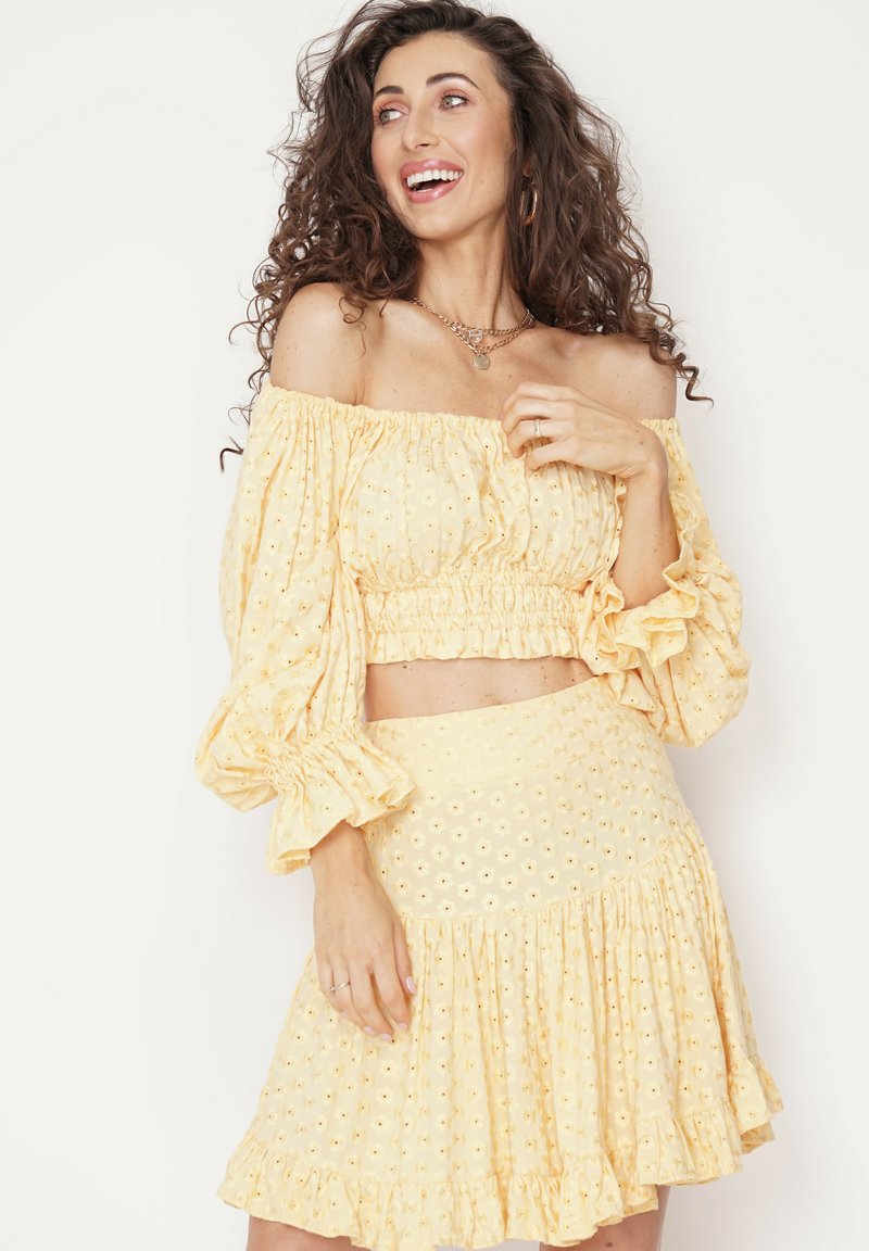 Żółty krótki top bez ramiączek z falbaniastymi rękawami, w zestawie z dopasowaną spódnicą w warstwy. Oba elementy mają haftowane detale w kwiaty.