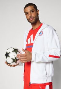 White zip-up jacket with red accents, worn over a red t-shirt featuring blue text and a logo. Holding a black and white soccer ball.