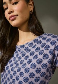 Lila bomullströja med ett blått blommönster, rund halsringning, korta ärmar och mjuk textur. Modellen ler och visar upp tröjan.