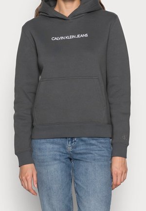 Personne portant un sweat à capuche Calvin Klein Jeans gris foncé avec une poche avant et un jean bleu clair, debout devant un fond uni.