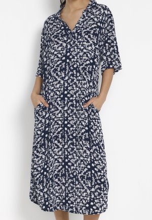 Woman wearing navy blue midi dress with white floral pattern, V-neck, short sleeves, and hands in side pockets.
