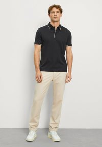 Schwarzes Poloshirt mit weißem Kragenrand, kombiniert mit beigen Hosen und weißen Sneakers. Der Stoff wirkt glatt und figurbetont.