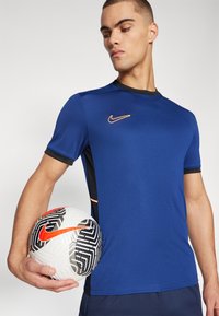 Blå Nike sportskjorte med sorte accenter og en orange logo, kombinert med en mønstrete hvit fotball med en fremtredende logo.