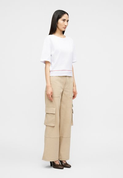 Γυναίκα που στέκεται πλευρικά, φοράει λευκό τοπ με κοντά μανίκια, μπεζ παντελόνι cargo με φαρδιές μπατζάκια και σκούρα καφέ παπούτσια με τακούνι.