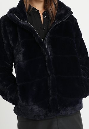 Giacca nera in ecopelliccia con collo alto e zip indossata sopra una camicia nera abbottonata, mani nelle tasche della giacca.