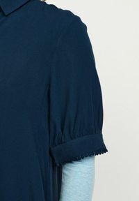 Tengerészkék póló rövid puffos ujjal, hajtott mandzsettával, pom-pom díszítéssel. Puha, könnyű szövettextúra.