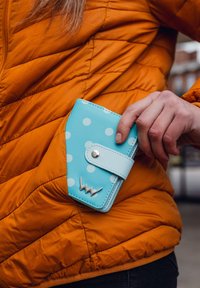 Hand holding light blue polka-dot wallet, partially inserted into pocket of orange quilted jacket.