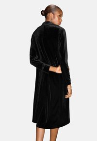 Abrigo largo de terciopelo negro con una textura suave, diseño sin cuello y mangas largas, con un corte recto y una silueta minimalista.