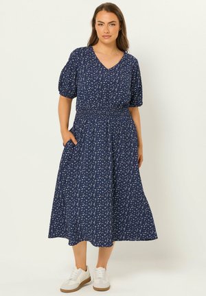 Kvinde iført en navyblå midikjole med hvidt blomsterprint, pufærmer, V-hals og hvide sneakers, stående med den ene hånd i en lomme.