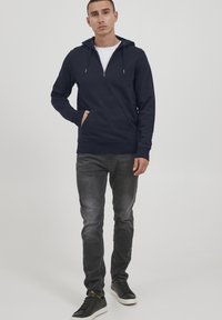 Mornarska zip-up kapuca z kenguru žepom, rebrastimi manšetami in robom. Povezana s sivimi kavbojkami in črnimi supergami, stoji ob enotni ozadju.
